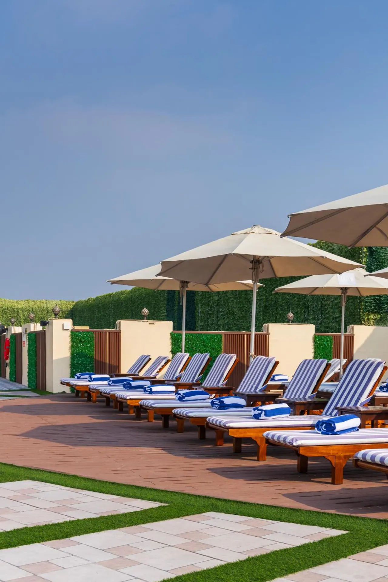 Rows of striped sun loungers with rolled blue towels and beige parasol umbrellas sit on a brick deck beside a green hedge under a clear blue sky.