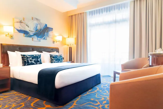 A bedroom with a king bed, blue accent pillows and a throw, two armchairs, and sheer-curtained floor-to-ceiling windows.
