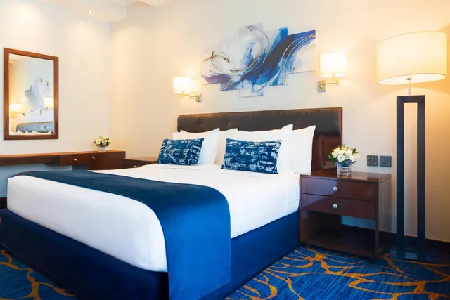 A bedroom with a made double bed with blue accents, a tall floor lamp, a bedside table with flowers, and a wall mirror.