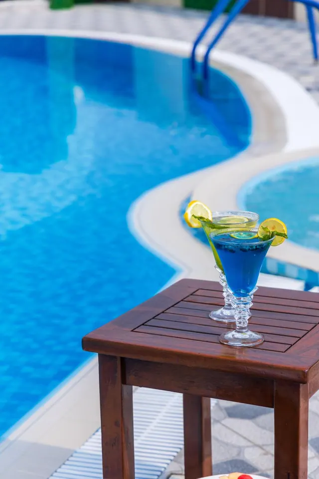 A blue cocktail garnished with lime and mint sits on a wooden side table beside a curved swimming pool.