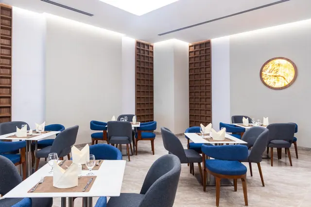 An empty restaurant dining room with white tables set with folded napkins, glasses and cutlery, blue and grey upholstered chairs, and wood-panelled wall niches.