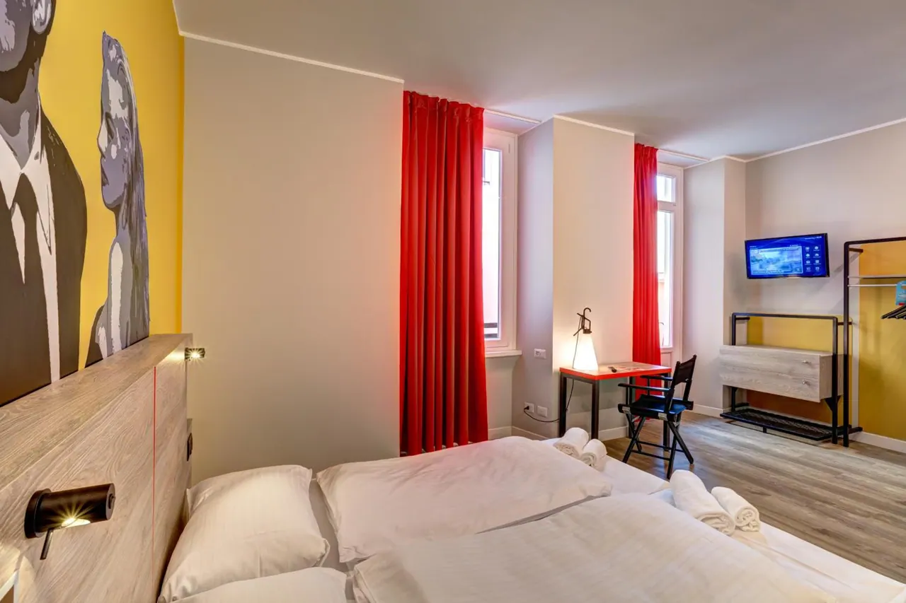 A bedroom with white bedding, a small red desk with a lamp and chair, red curtains, a wall-mounted television and an open clothing rack.