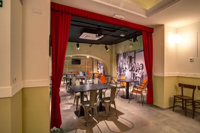 A restaurant dining room framed by red curtains, with dark tables, metal and orange chairs, and a large black-and-white photo mural on the wall.