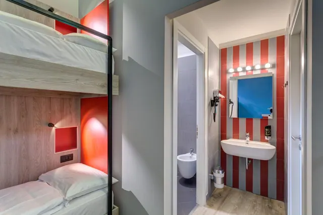 Bunk beds with white bedding sit beside a bathroom with a red and white striped wall, a sink with a lit mirror, and a bidet visible through the open door.