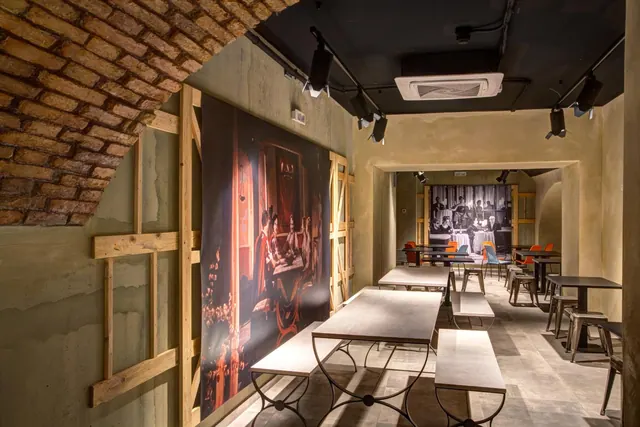 A dining room with long metal-framed tables, benches and stools, a large printed artwork on the left wall and a brick arch overhead.