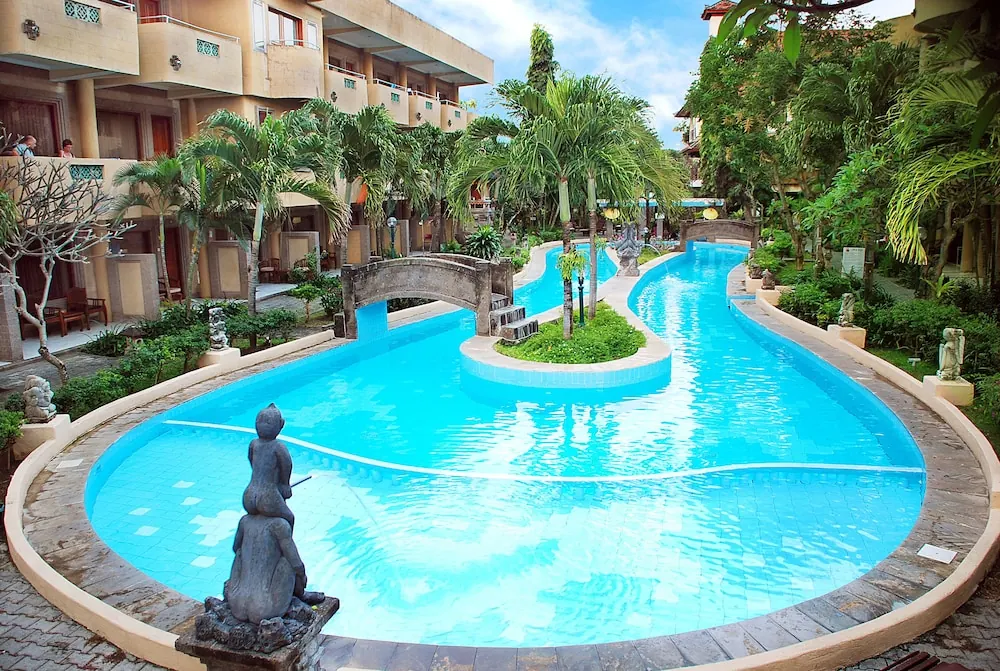 A winding lazy river pool lined with palm trees and garden planting, with statues on the deck, a small stone bridge, and hotel balconies behind.
