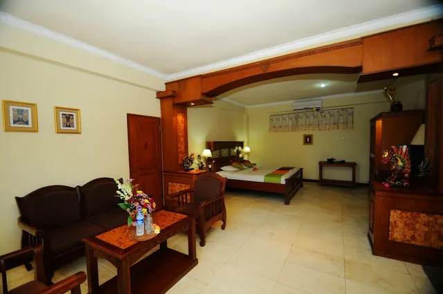 A hotel room with a dark wooden sofa and carved coffee table holding a flower arrangement in the foreground, with a double bed, wall-mounted air conditioner and wooden wardrobe in the background.
