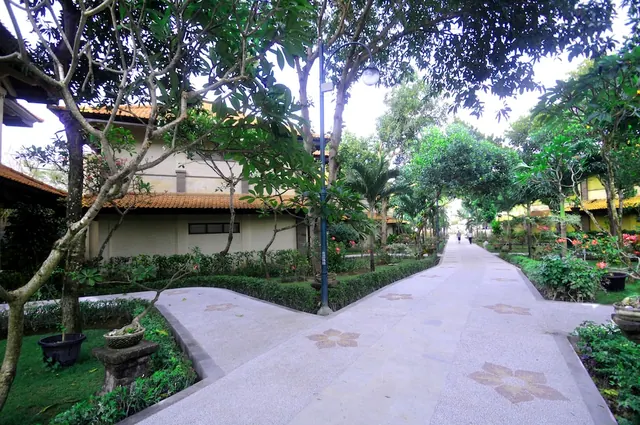 A tree-lined paved pathway with decorative floor tiles runs between low villas and garden beds, with a lamp post on the left and two people far down the path.