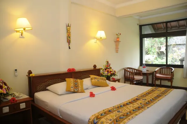 A bedroom with a king bed dressed with yellow cushions, red hibiscus flowers and a batik runner, with wall lamps, a flower arrangement and a window looking onto green foliage.