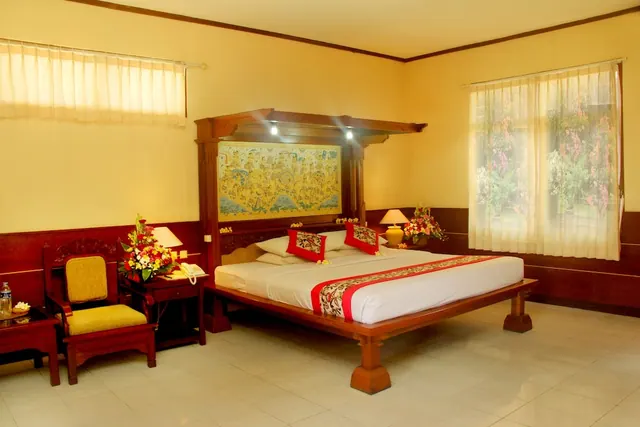 A bedroom with a carved wooden bed under a painted panel, a yellow armchair, flower arrangements, and a window with sheer curtains looking onto greenery and flowering plants.