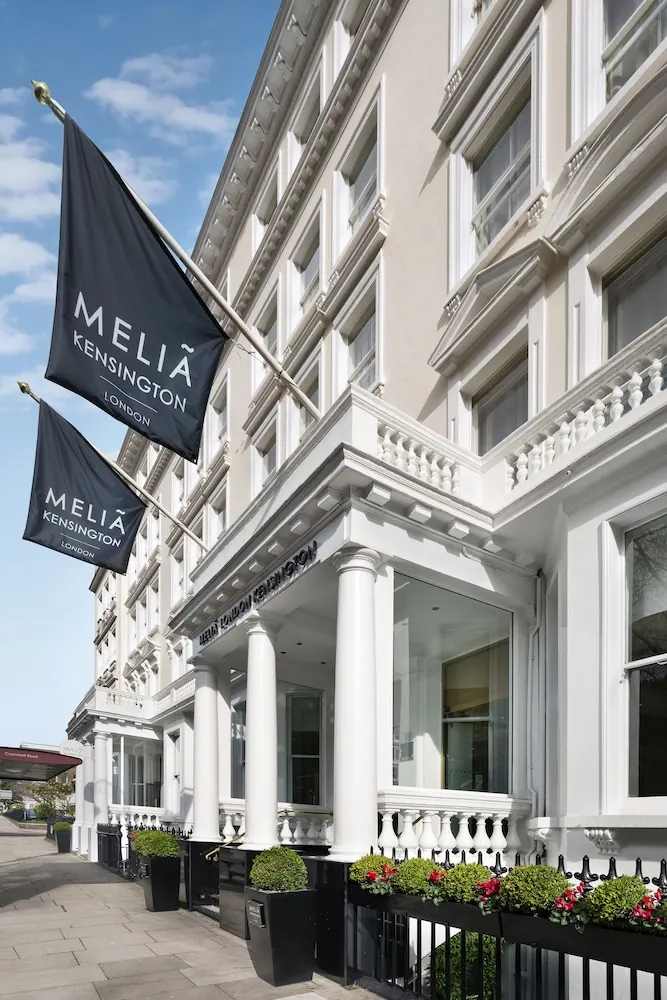 Exterior of the Meliá Kensington hotel showing a white stucco facade, columns, street-level planters and two navy flags under a blue sky.