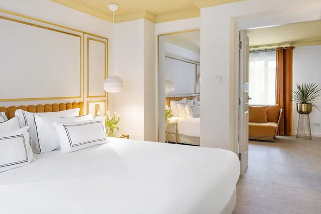A made-up white bed with a tufted mustard headboard and gold-trimmed wall panels, with a second bedroom and a seating area with a mustard sofa visible through open doorways.