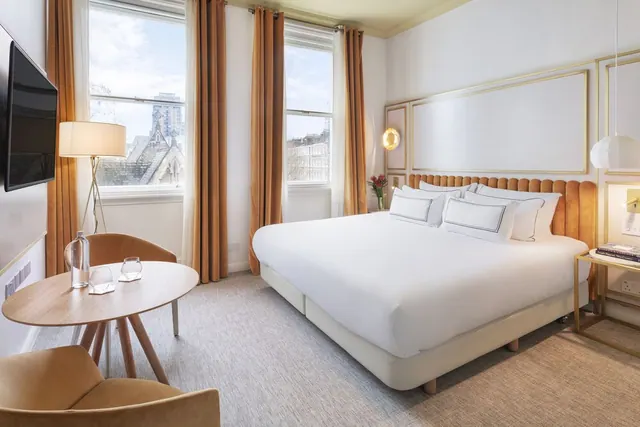 A bedroom with a made-up king bed, a wall-mounted television, a small round table with two armchairs, and tall windows looking out over buildings.