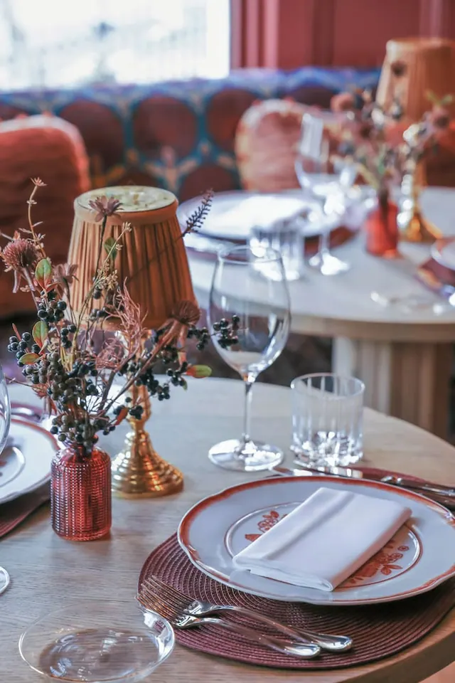 A restaurant table is set with a patterned plate, folded white napkin, cutlery, wine glasses, a tumbler and dried flower arrangements, with a banquette seat behind.