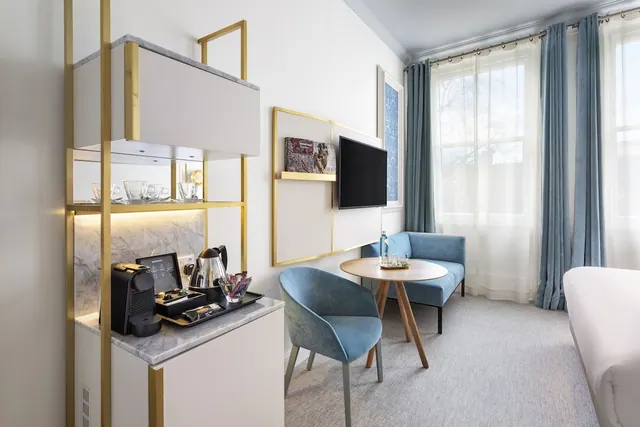 A hotel room seating area with blue armchairs and a sofa, a round wooden table, a wall-mounted television and a coffee station with a kettle and coffee machine.