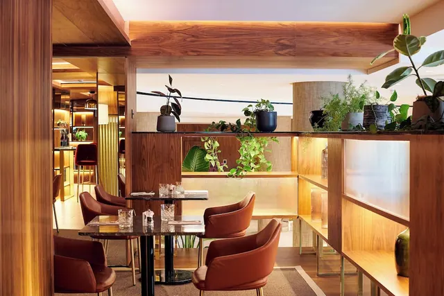 A restaurant dining area with wood panelling, leather armchairs at marble-topped tables, and plants on backlit shelving.