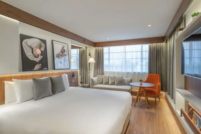 A staged hotel bedroom with a king bed, a grey sofa, an orange armchair beside a round table, and a wall-mounted television.