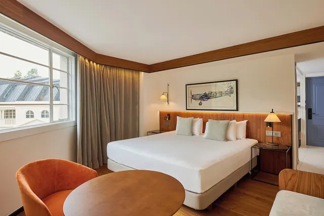 A hotel bedroom with a made king bed, a wooden headboard, an orange armchair and a round wooden table beside a window looking onto a neighbouring building.