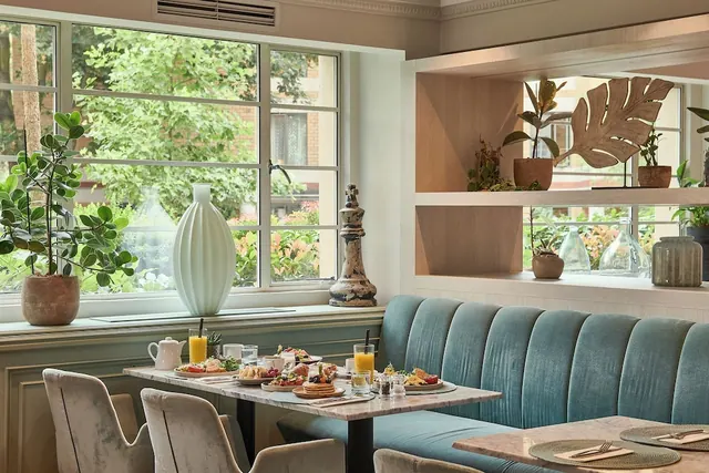 A dining room with teal banquette seating, a table set with breakfast dishes and juice, and large windows looking onto trees and plants outside.