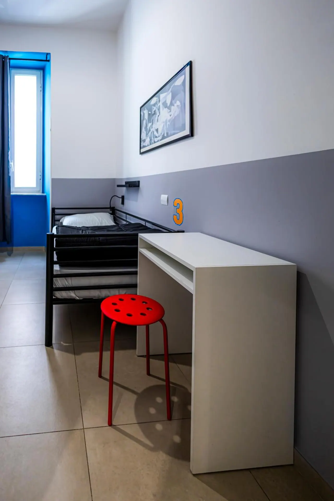 A small bedroom with a black metal bunk bed, a white desk, a red stool and a framed black and white print on the wall.