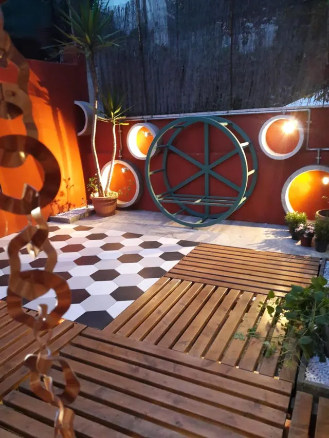 A dusk courtyard with a hexagonal tiled floor, wooden decking, a red wall with round lit niches, a teal wheel-shaped ornament and a potted plant.