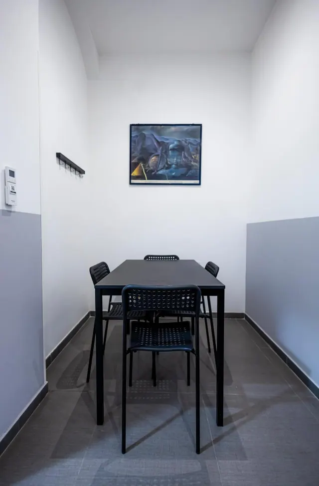 A small room with a black table and four black chairs, a framed painting on the white wall, and a wall-mounted intercom panel beside a coat hook rail.