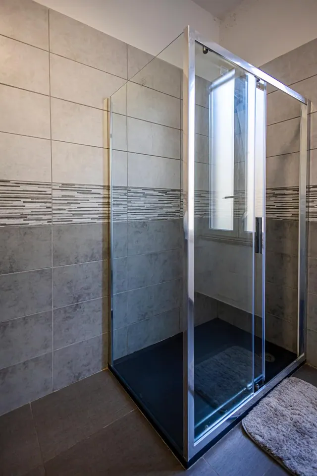 A glass-enclosed walk-in shower with grey wall tiles, a dark shower tray and a grey bath mat on the tiled floor.