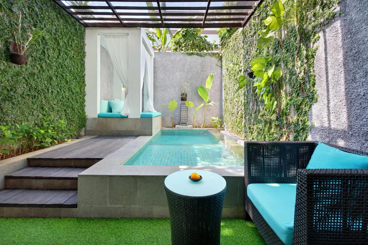 A private plunge pool with turquoise tiles sits in a walled courtyard with ivy-covered walls, a pergola roof, a daybed with curtains and a wicker armchair with turquoise cushions.