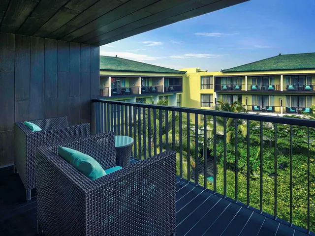 A private balcony with two wicker armchairs with teal cushions, a round side table and a railing overlooking trees and a neighbouring hotel building.