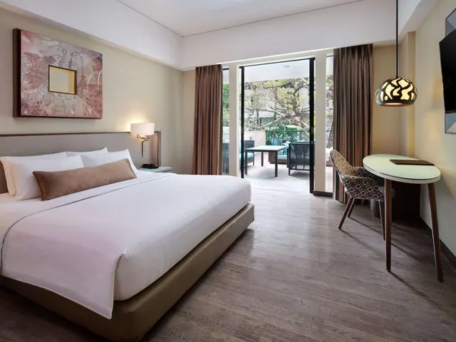 A king bed, a round work desk and glass sliding doors opening onto a balcony with furniture and trees are visible in this bedroom.