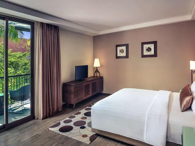 A bedroom with a made king bed, a wall-mounted TV on a wooden sideboard, and sliding glass doors opening onto a balcony with tropical greenery beyond.
