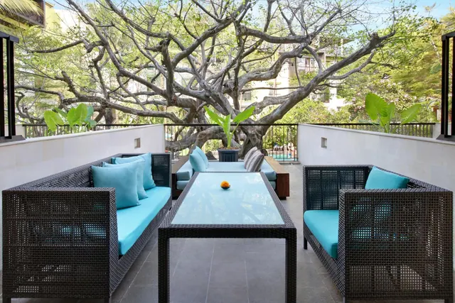 A terrace with dark wicker sofas, turquoise cushions and a glass-top coffee table, with a large bare-branched tree and greenery beyond the railing.