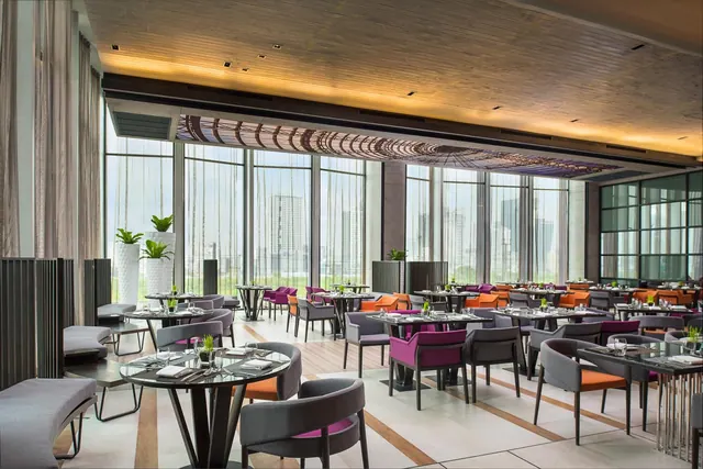 A restaurant dining room with rows of tables and upholstered chairs in grey, purple and orange, beside tall windows looking out onto high-rise buildings.