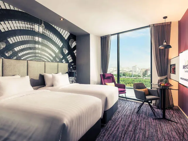 A bedroom with two single beds, a large arched-roof mural on the wall, purple and grey armchairs, and floor-to-ceiling windows looking out over a city.
