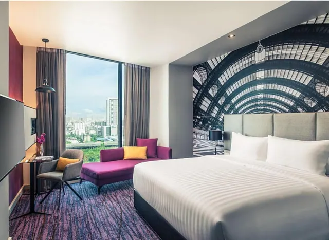 A hotel bedroom with a large bed, a purple chaise, a desk and chair, and a tall window overlooking city buildings.