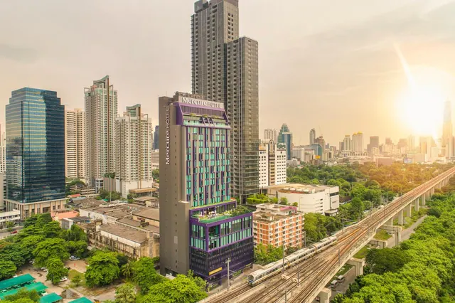 Elevated view of the Mercure Hotel tower in Bangkok, with surrounding high-rise buildings and an elevated rail line at sunset.