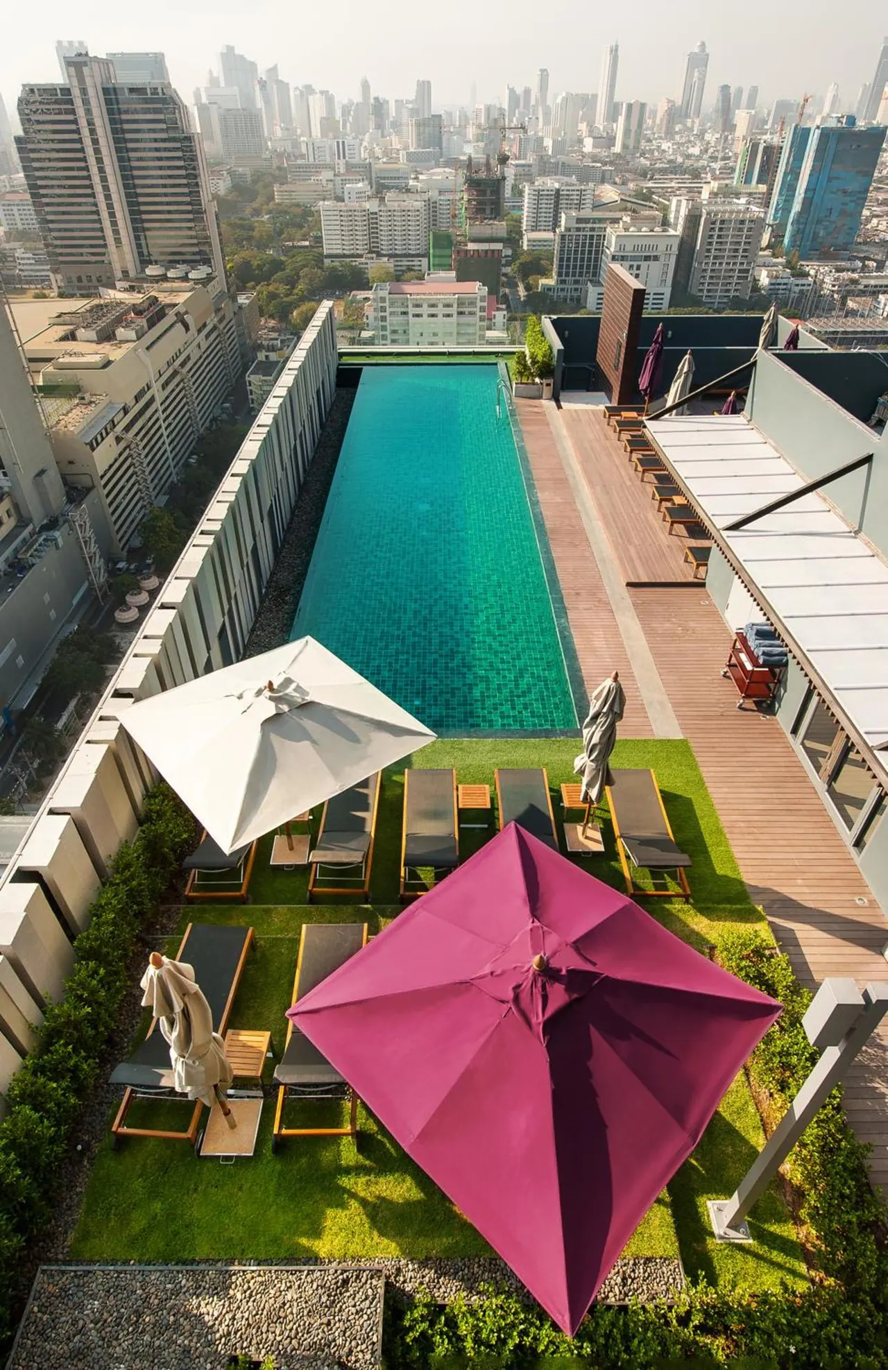 Overhead view of a long rooftop lap pool with sun loungers, umbrellas and a hazy city skyline in the background.