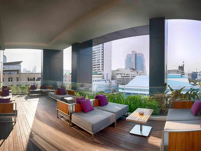 A covered rooftop terrace with grey sofas, purple cushions and a wooden deck overlooks city towers under a hazy sky.