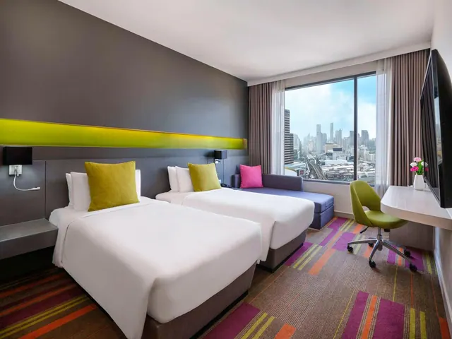 A hotel room with two beds, a purple sofa, a green desk chair, a wall-mounted television and a window overlooking a city skyline.
