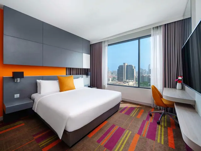 A king bed with orange accent cushions sits against a grey panelled wall, with a desk and wall-mounted television on the right and a window overlooking city towers.