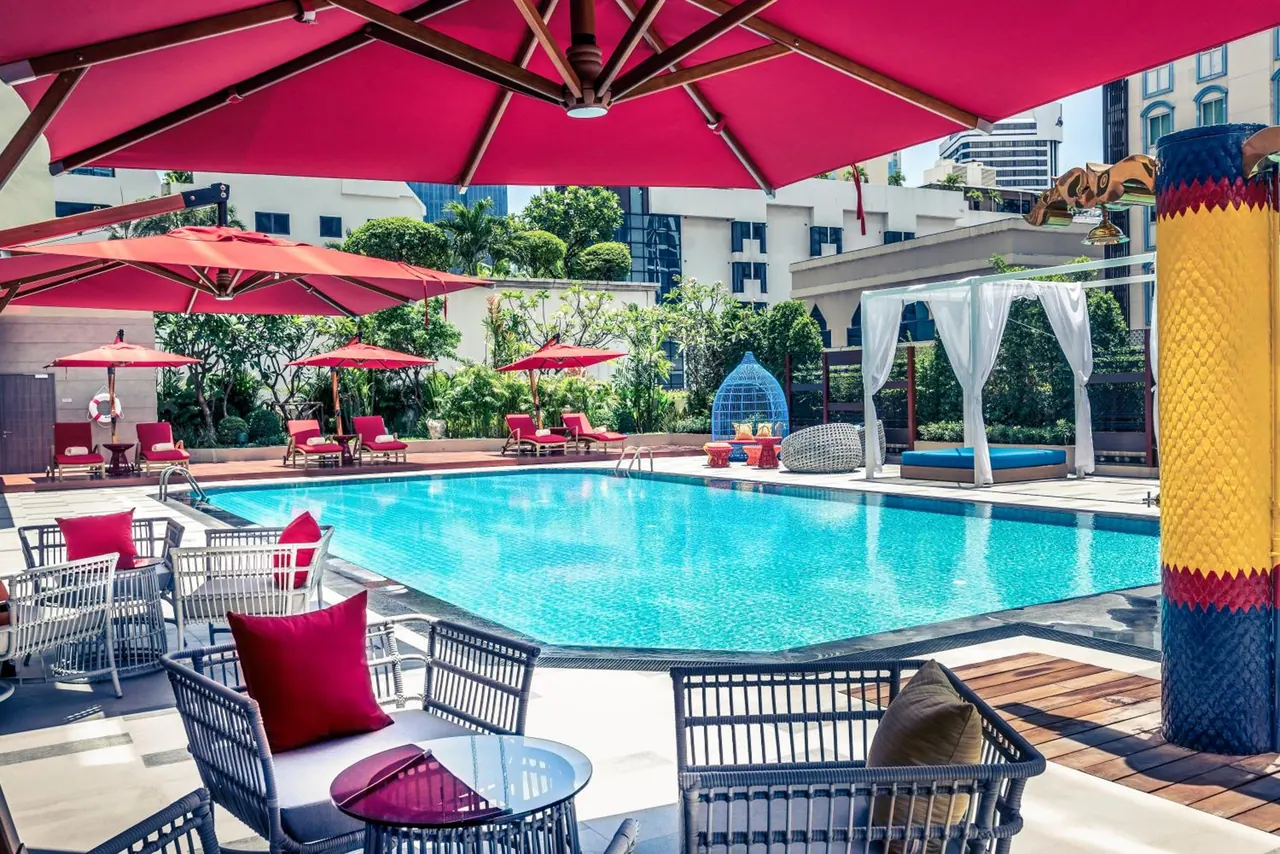 A large swimming pool with red umbrellas, rattan chairs, sun loungers and a white canopy daybed beside it.