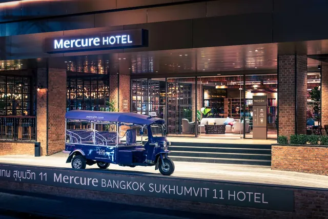 A blue tuk-tuk is parked in front of the Mercure Hotel entrance at night, with the lit lobby and a seating area visible behind it.