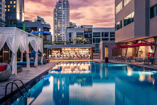 The hotel pool at dusk, with a lit poolside bar, white canopied cabanas, red umbrellas and city towers behind.