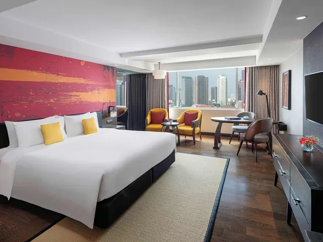 A king bed with yellow and white bedding sits beside a red painted accent wall, with two yellow armchairs and a round table by a window overlooking city high-rises.
