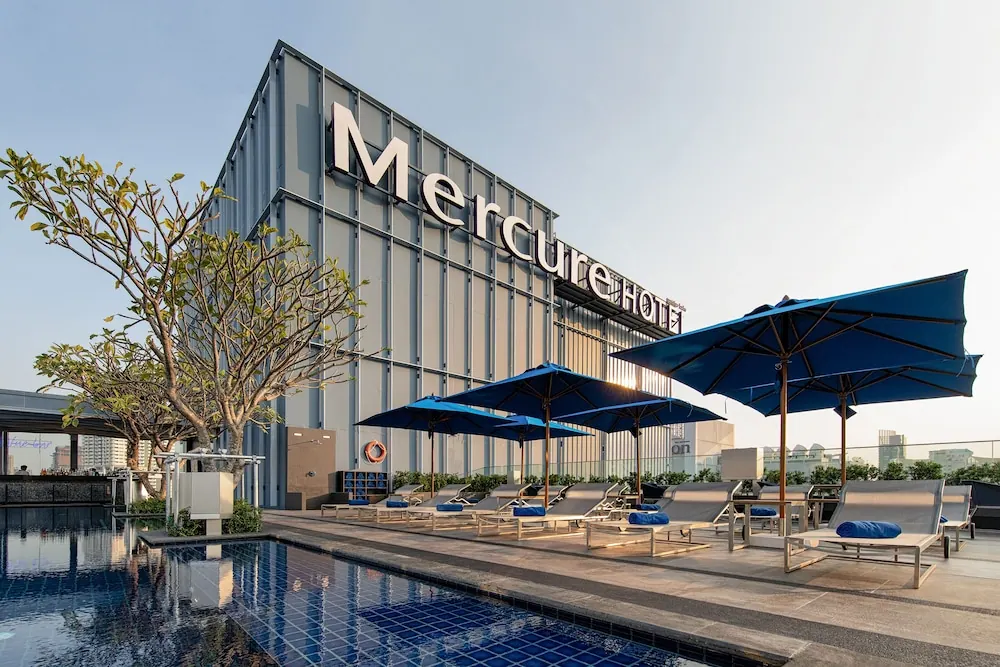 A rooftop pool with blue umbrellas and sun loungers in front of a building bearing the Mercure sign.