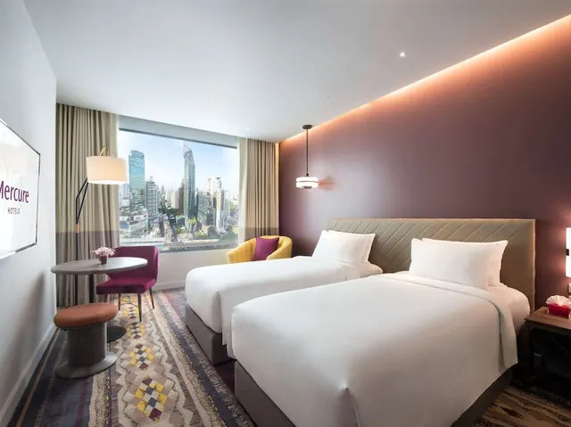 A twin bedroom with two white beds, a yellow armchair, a round table with pink chairs, and a large window overlooking city skyline buildings.