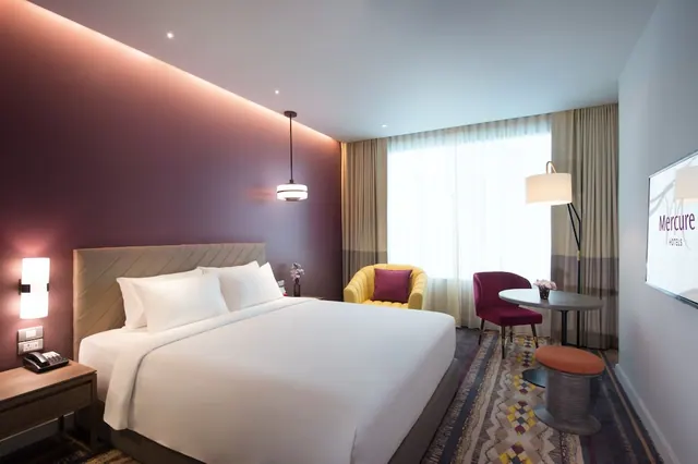 A hotel bedroom with a large white bed, a yellow armchair and a red chair at a round table, and a wall-mounted television.