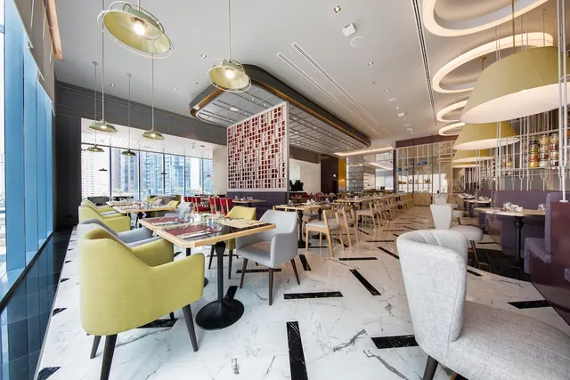 A bright restaurant dining room with yellow and grey armchairs at tables, pendant lamps, marble floors, and windows showing city towers.