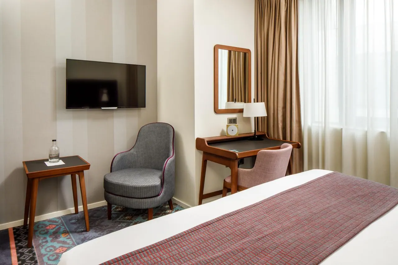 A hotel bedroom with a wall-mounted television, a grey armchair beside a small side table, a wooden desk with a chair and mirror, and a made bed in the foreground.