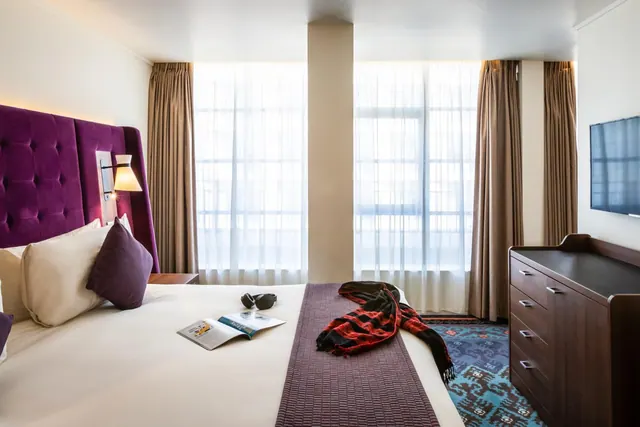 A bedroom with a double bed under a tall purple tufted headboard, a magazine and sunglasses on the bed, a wooden dresser and a wall-mounted television, with sheer-curtained windows behind.